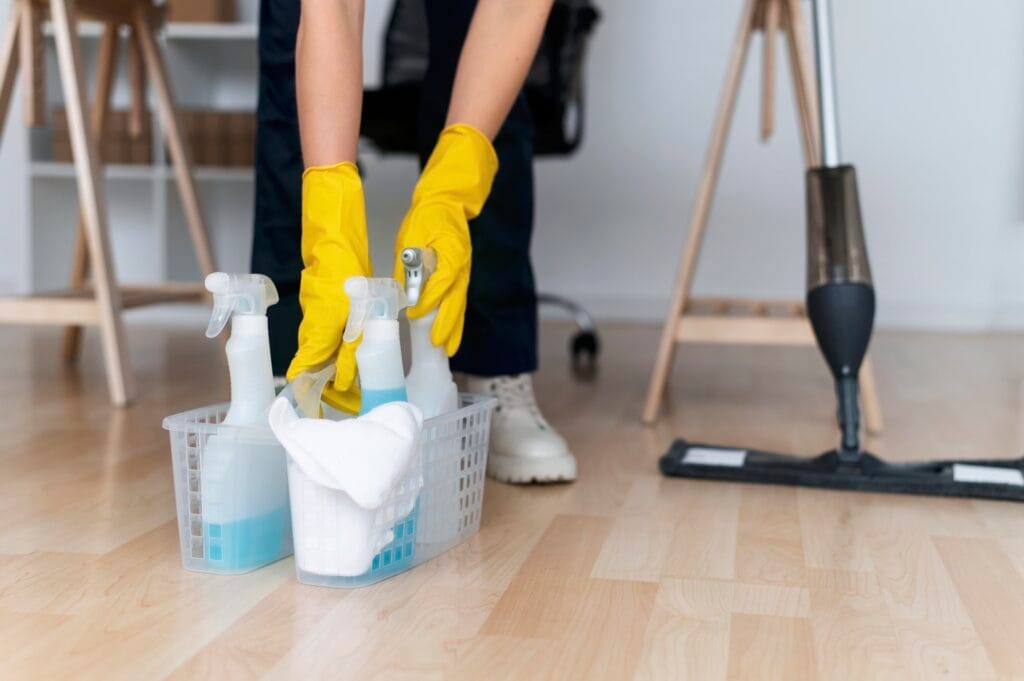Person wearing yellow gloves cleaning spray bottles in a plastic basket on a wooden floor.