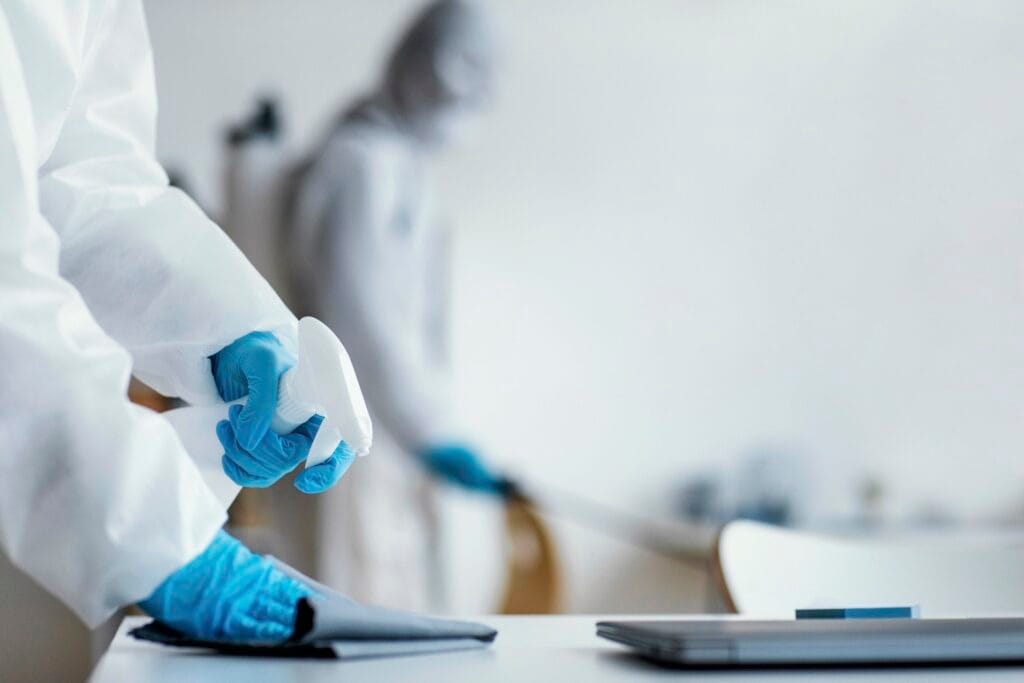 Person in protective gear holding a spray bottle, with another person blurred in the background.