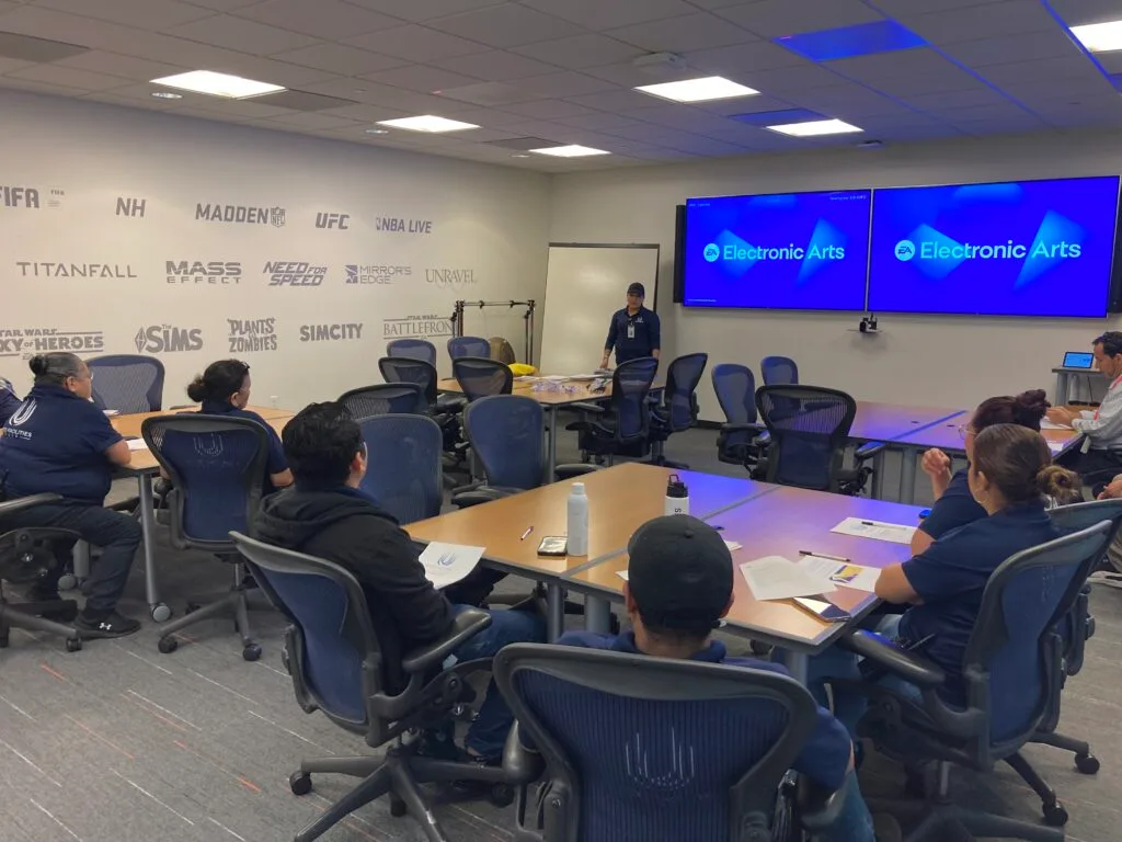 People seated around a conference table watching a presenter in a room with EA and video game logos on the wall.