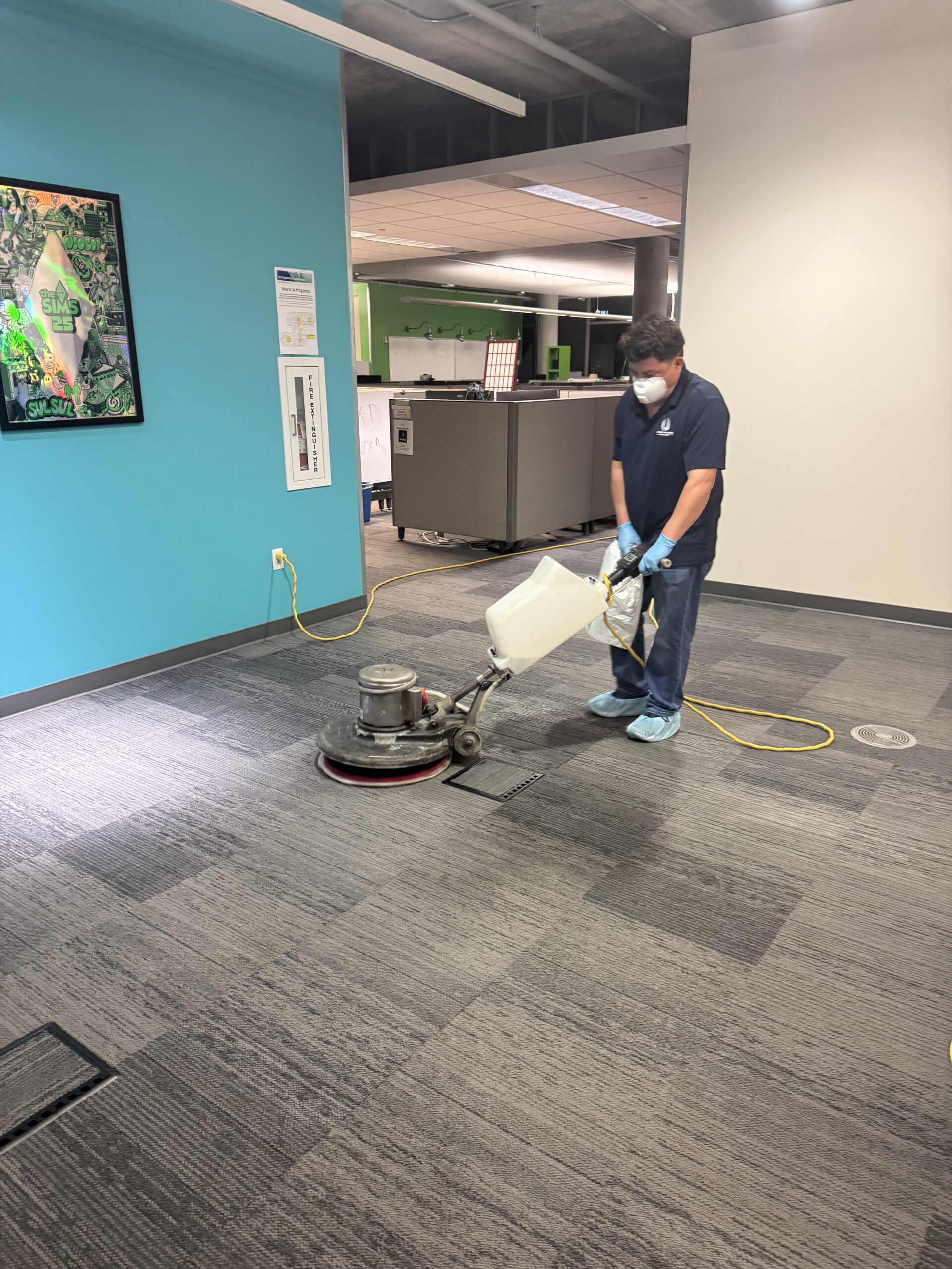Person wearing a mask and gloves operating a floor cleaning machine in an office space.