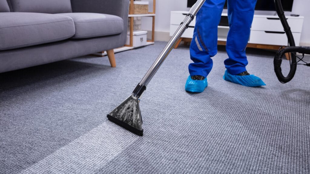 Person in blue protective shoe covers vacuuming a gray carpet in a living room.