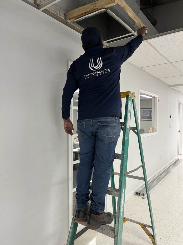 Worker on ladder installing ceiling tiles in an office space, wearing a navy jacket with United Facilities Group logo.