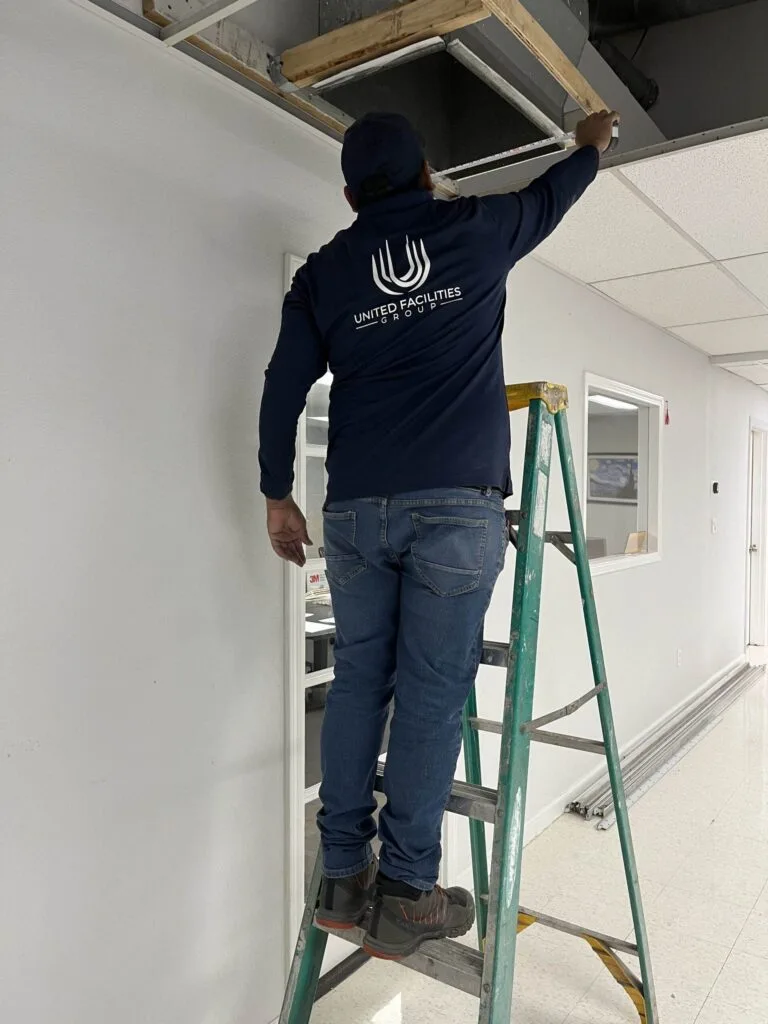 Worker on ladder installing ceiling tiles in an office space, wearing a navy jacket with United Facilities Group logo.