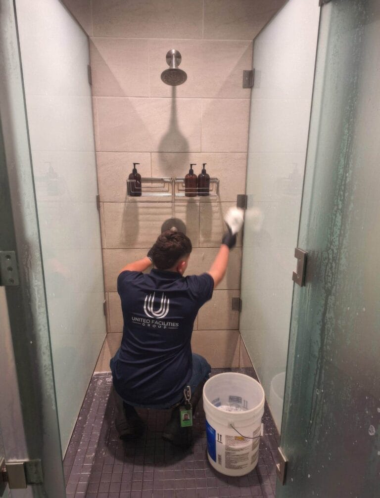Worker cleaning shower glass with a squeegee, kneeling inside a tiled shower stall.