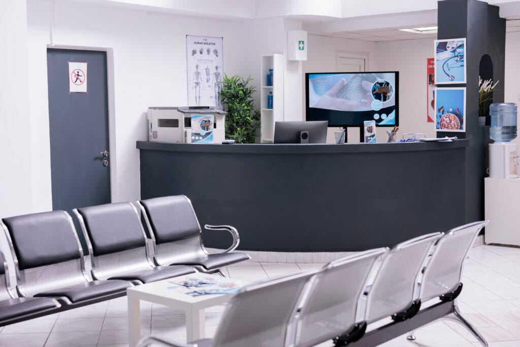 Empty waiting area with black and silver chairs in front of a dark reception desk in a medical office.