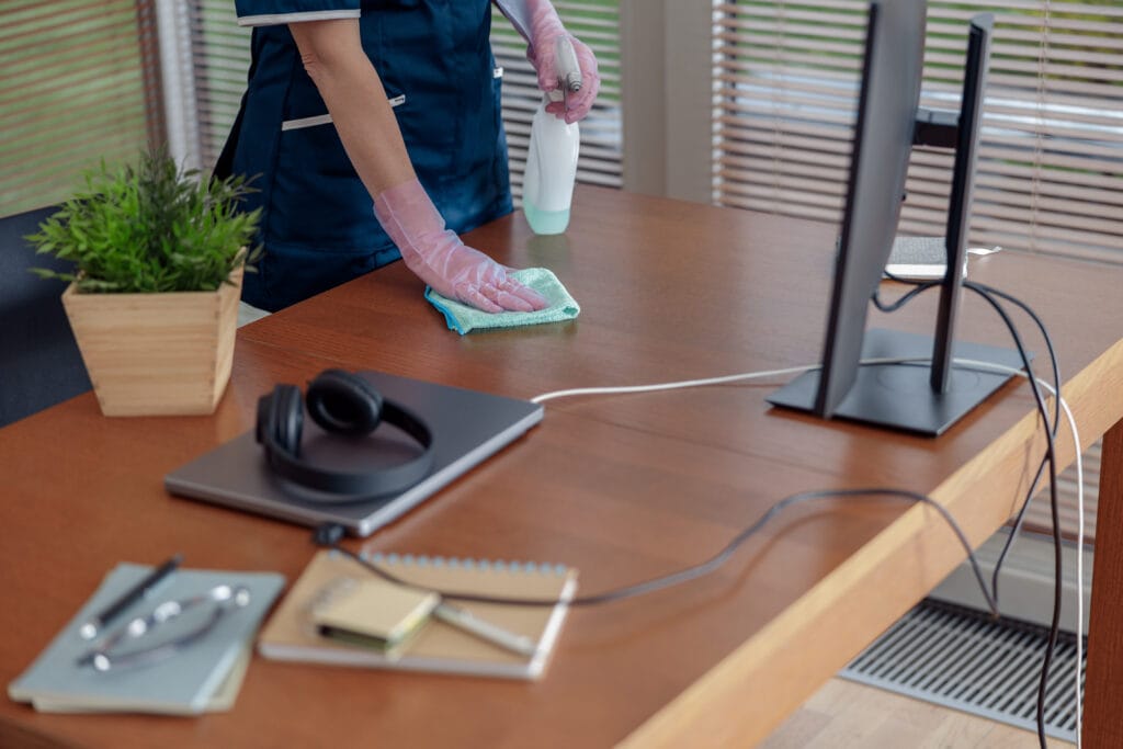 Person cleaning a wooden desk with a spray bottle and cloth, wearing pink gloves.