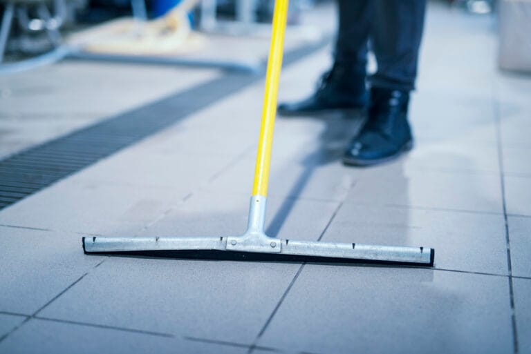Person wearing black shoes and pants sweeping tiled floor with a yellow-handled broom.