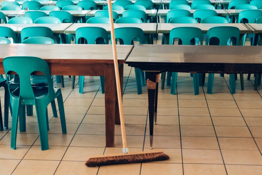 A broom leaning against a wooden table in front of multiple teal plastic chairs and tables.