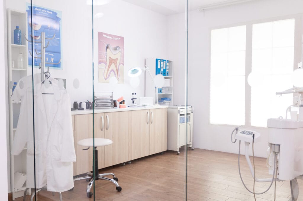 Bright dental clinic with posters, a white coat on a hanger, and dental equipment near windows.