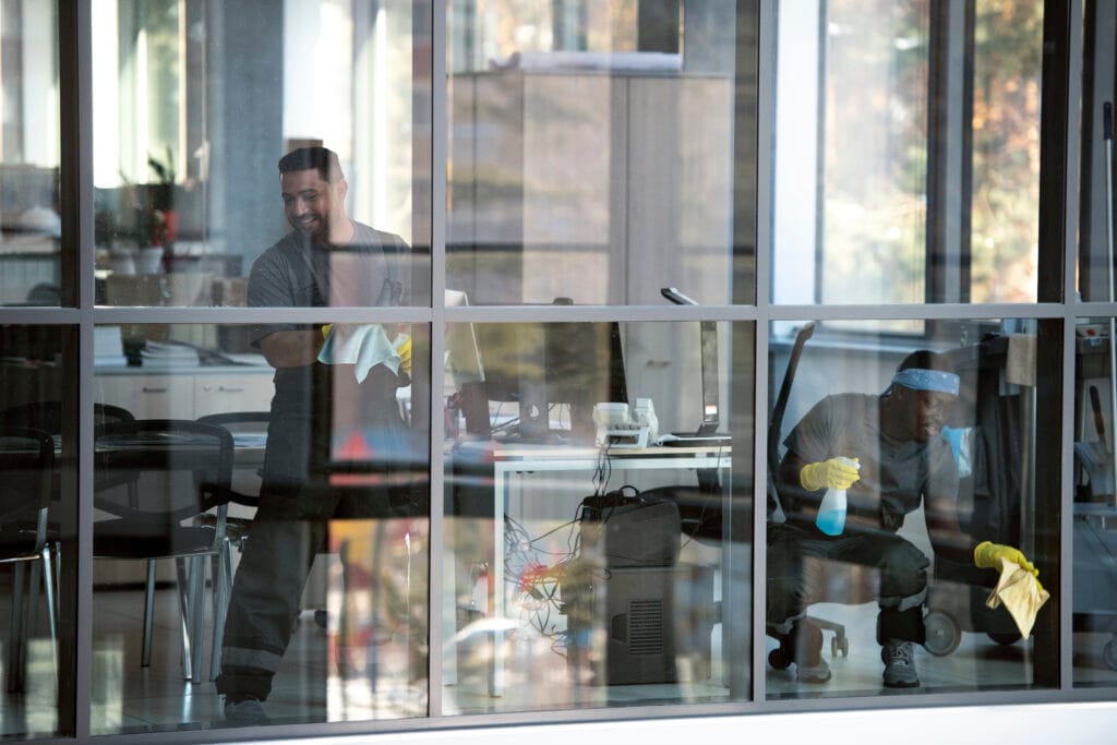 Two men cleaning an office, one standing with a cloth and the other kneeling with a spray bottle.