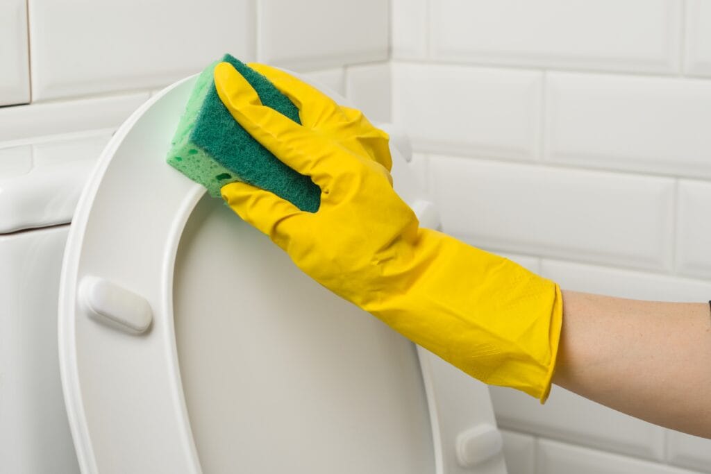 Hand wearing a yellow glove cleaning a white toilet with a green sponge.