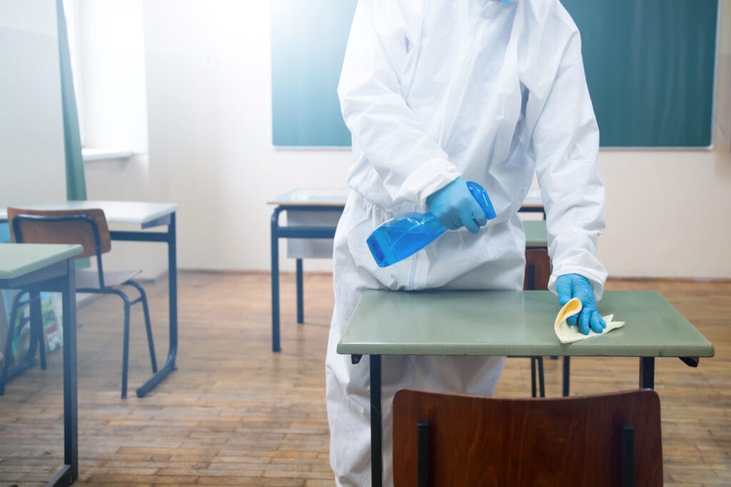 Person in protective suit disinfecting classroom desk with spray and cloth.