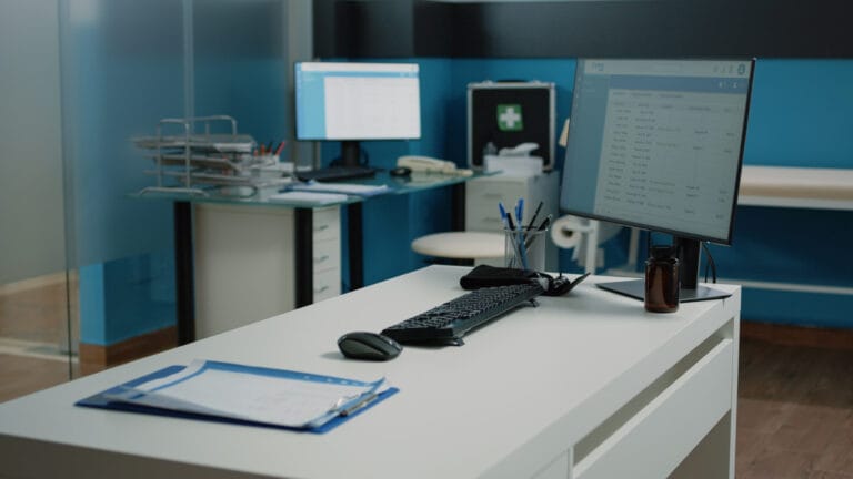 Desk with a computer monitor, keyboard, mouse, pen holder, and a clipboard in an office.