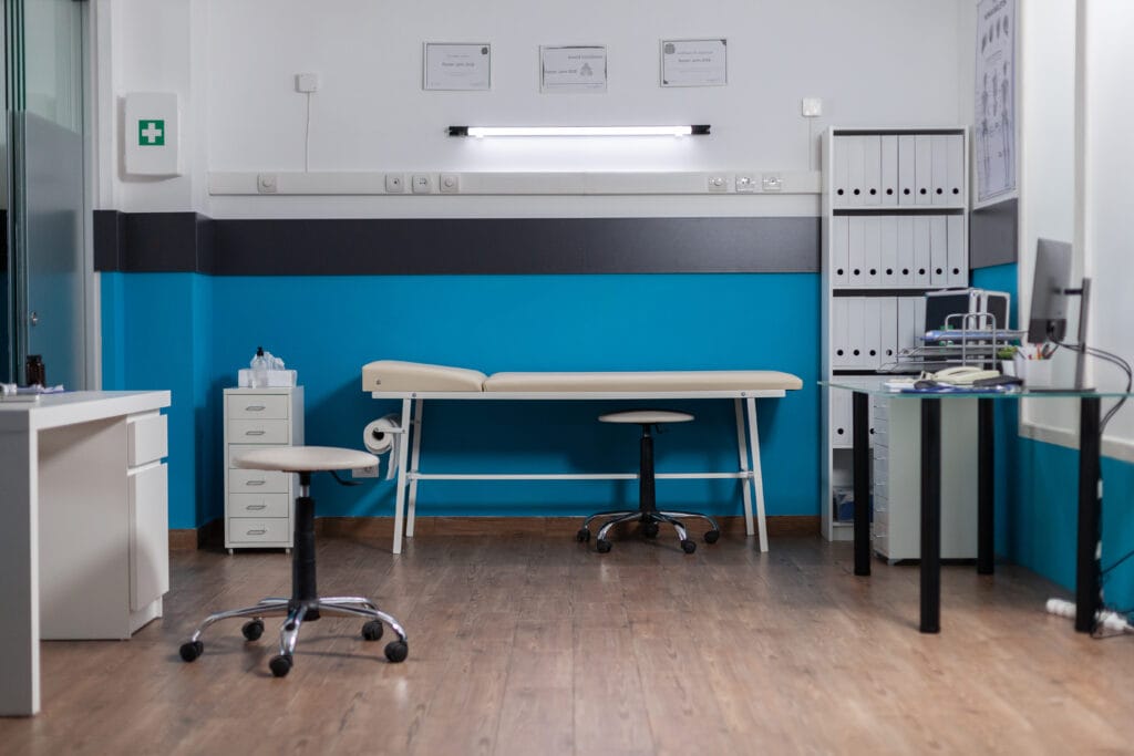 Empty medical examination room with a beige examination table, chairs, and medical supplies.
