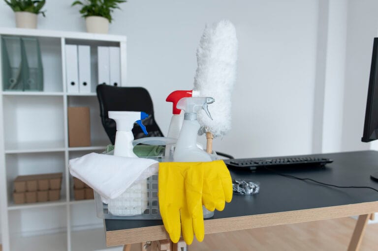 Cleaning supplies, including spray bottles, yellow gloves, and a duster, on a desk.