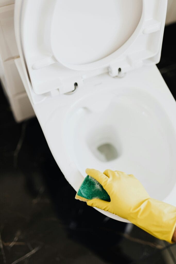 Person wearing yellow gloves cleaning a white toilet with a green sponge.