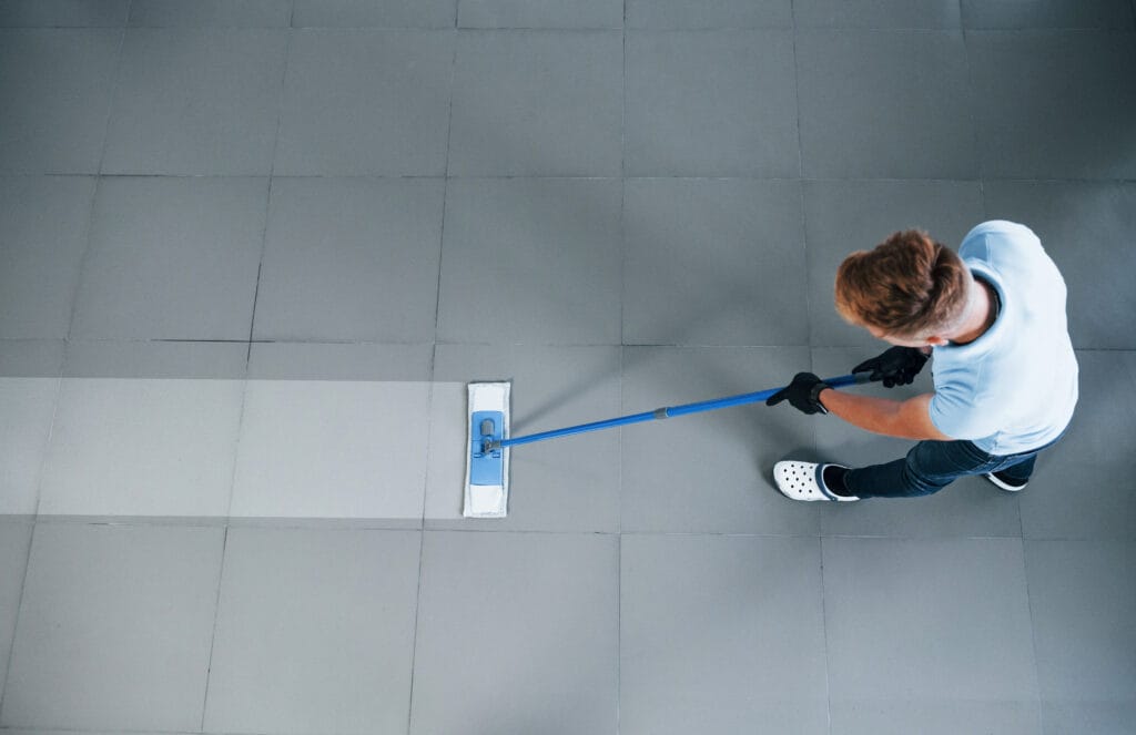Person mopping gray tiled floor, wearing white shirt, black gloves, and Crocs.