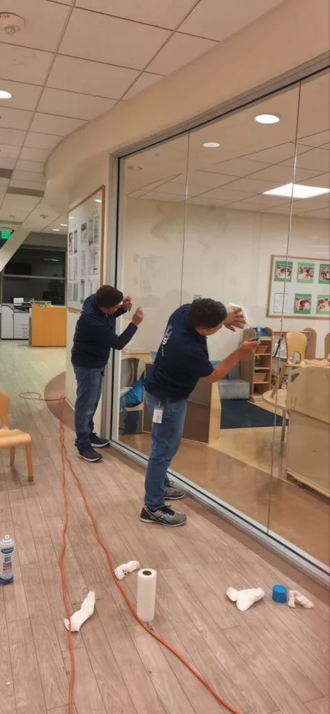 Two people cleaning glass windows inside a room with wooden floors, using cloths and spray bottles.