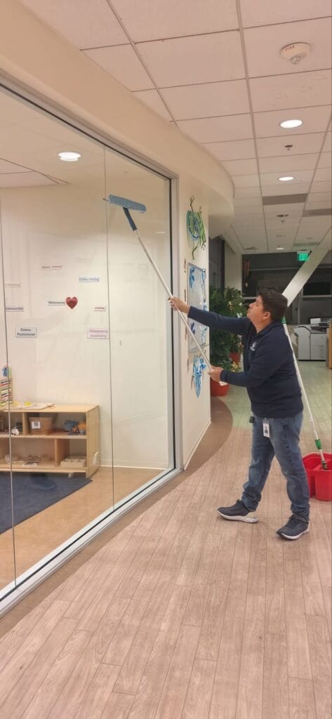 Person cleaning glass wall with a long-handled squeegee in an office corridor.