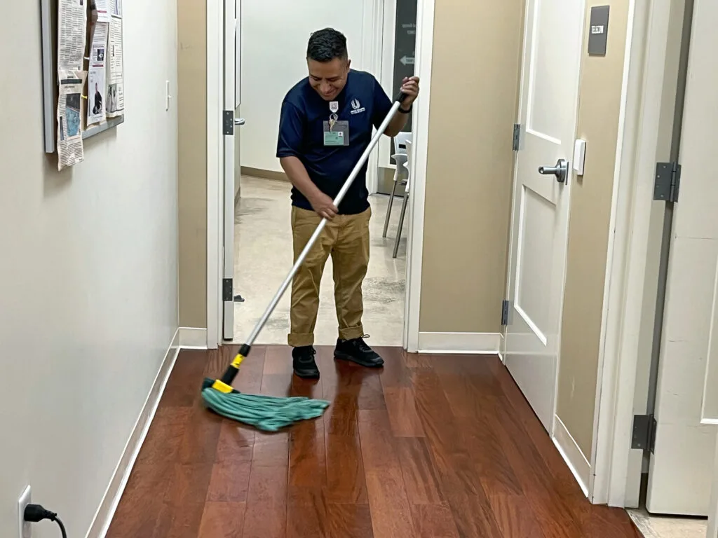 Person cleaning the floor with a mop in a hallway near closed doors and a bulletin board.