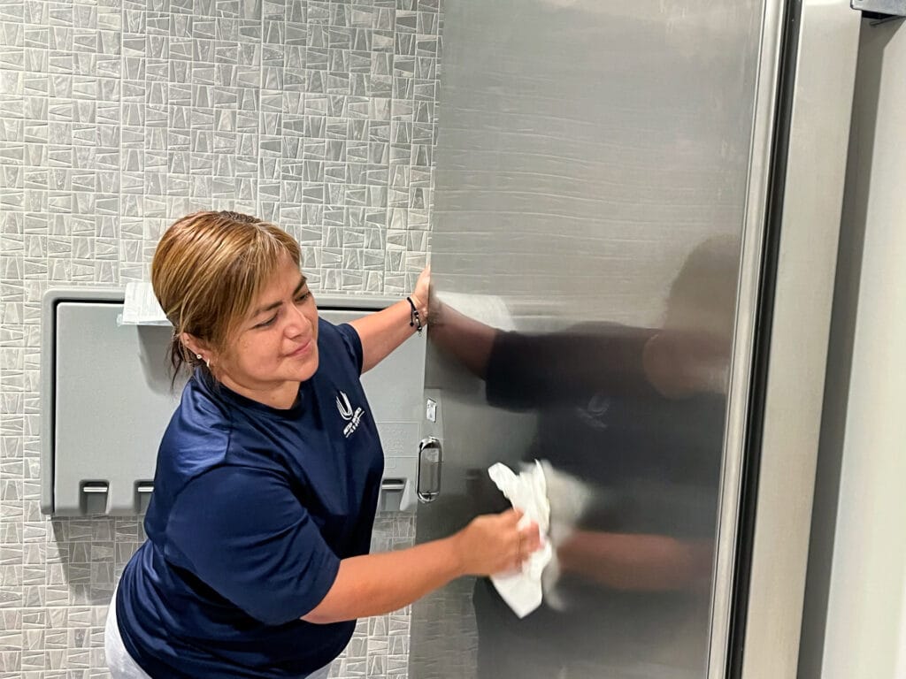 Woman cleaning a stainless steel surface in a tiled restroom.