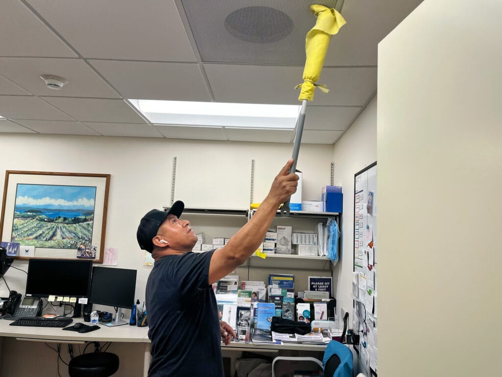 Person in a black cap and shirt using a long-handled duster to clean the ceiling.