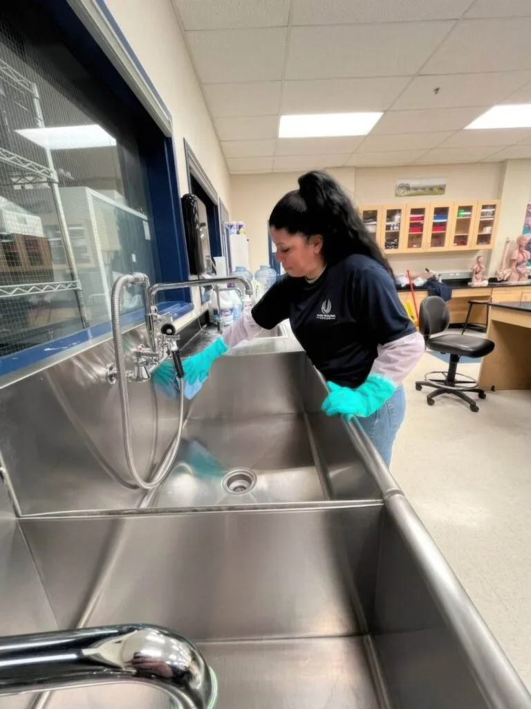 Woman cleaning a stainless steel sink in a laboratory or medical setting.