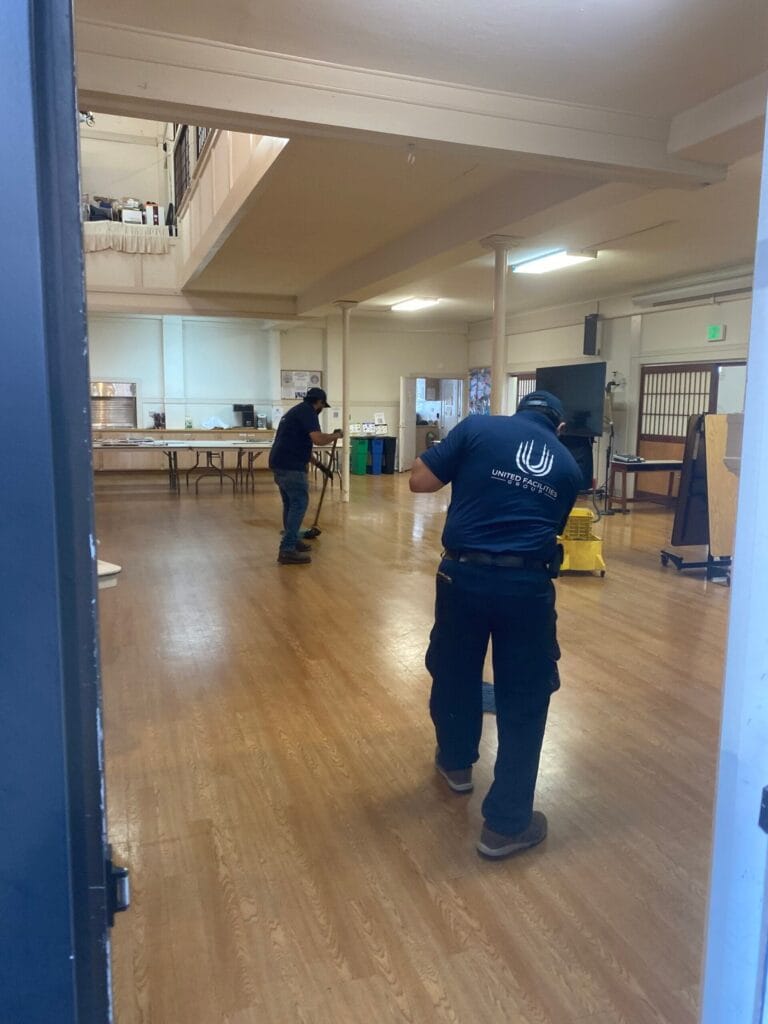 Two workers cleaning a large, empty room with wooden floors and high ceilings.