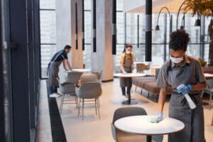 Three staff members wearing masks and gloves disinfect tables in a modern, high-rise restaurant.